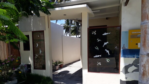 A building entrance with a glass door displaying fish, a small garden with plants, and a pathway leading to the entrance. - Hospedaje Mother Fanny (Puerto Villamil)