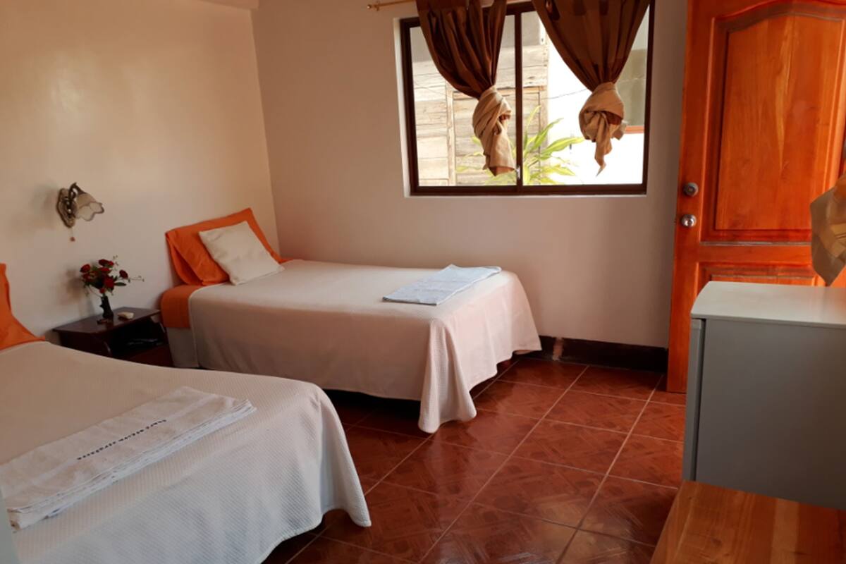 A hotel room with two beds, a window with curtains, a wooden door, and a tiled floor.