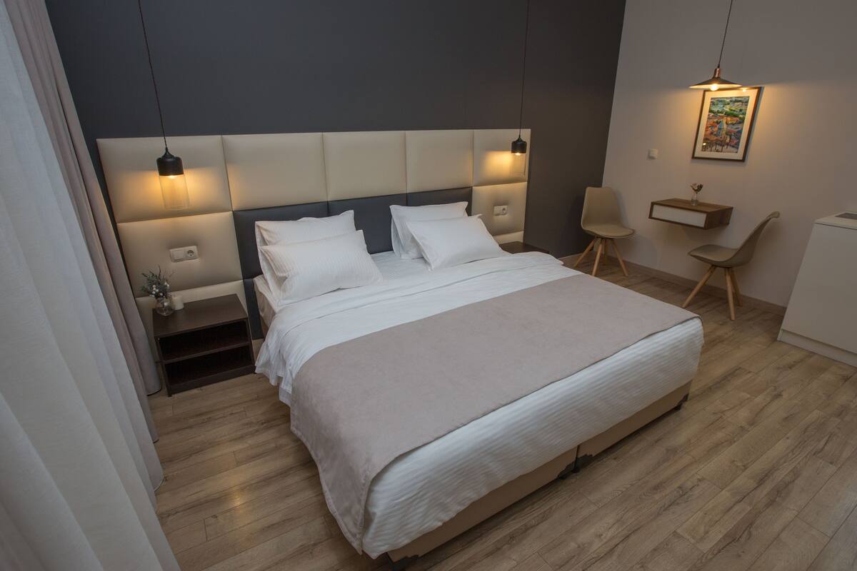 A modern hotel room with a large bed, a bedside table, a chair, and a desk with a lamp.