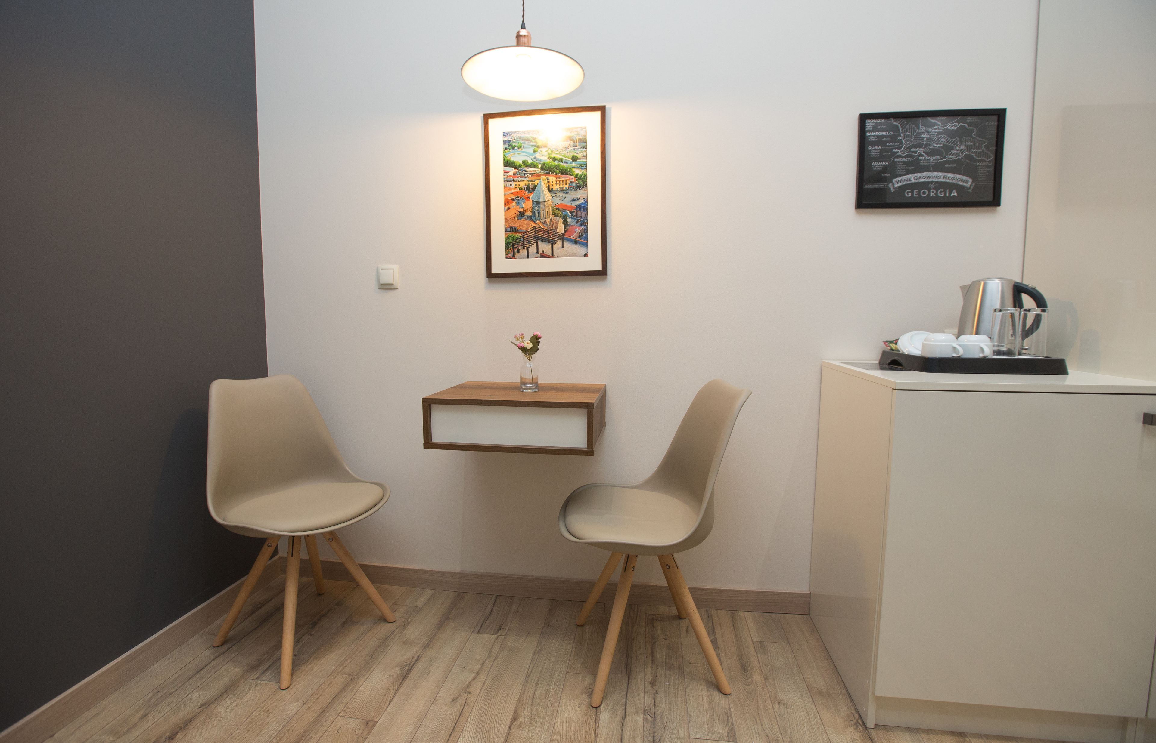 Una habitación con dos sillas, una mesita con un florero, un mapa colgado en la pared y un mueble con una tetera y tazas.