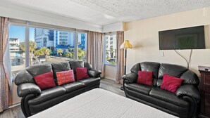 A modern living room with two black leather sofas, a flat-screen TV, and a large window offering a city view. - Ocean 5 Hotel (Myrtle Beach)