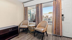 A room with two chairs, a small round table, and a dark wooden cabinet. A window with patterned curtains is visible, and a white door is on the right. - Ocean 5 Hotel (Myrtle Beach)