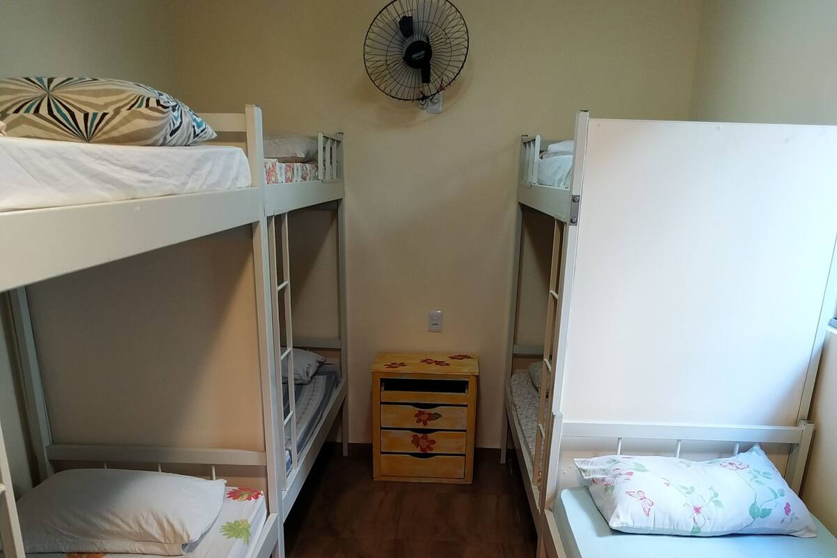A dormitory room with bunk beds, a wooden chest of drawers, and a wall-mounted fan.