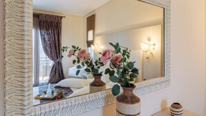 A bedroom with a large mirror, two vases with flowers, a bedside table, and a window with curtains. - Grand View Villas (Samos)
