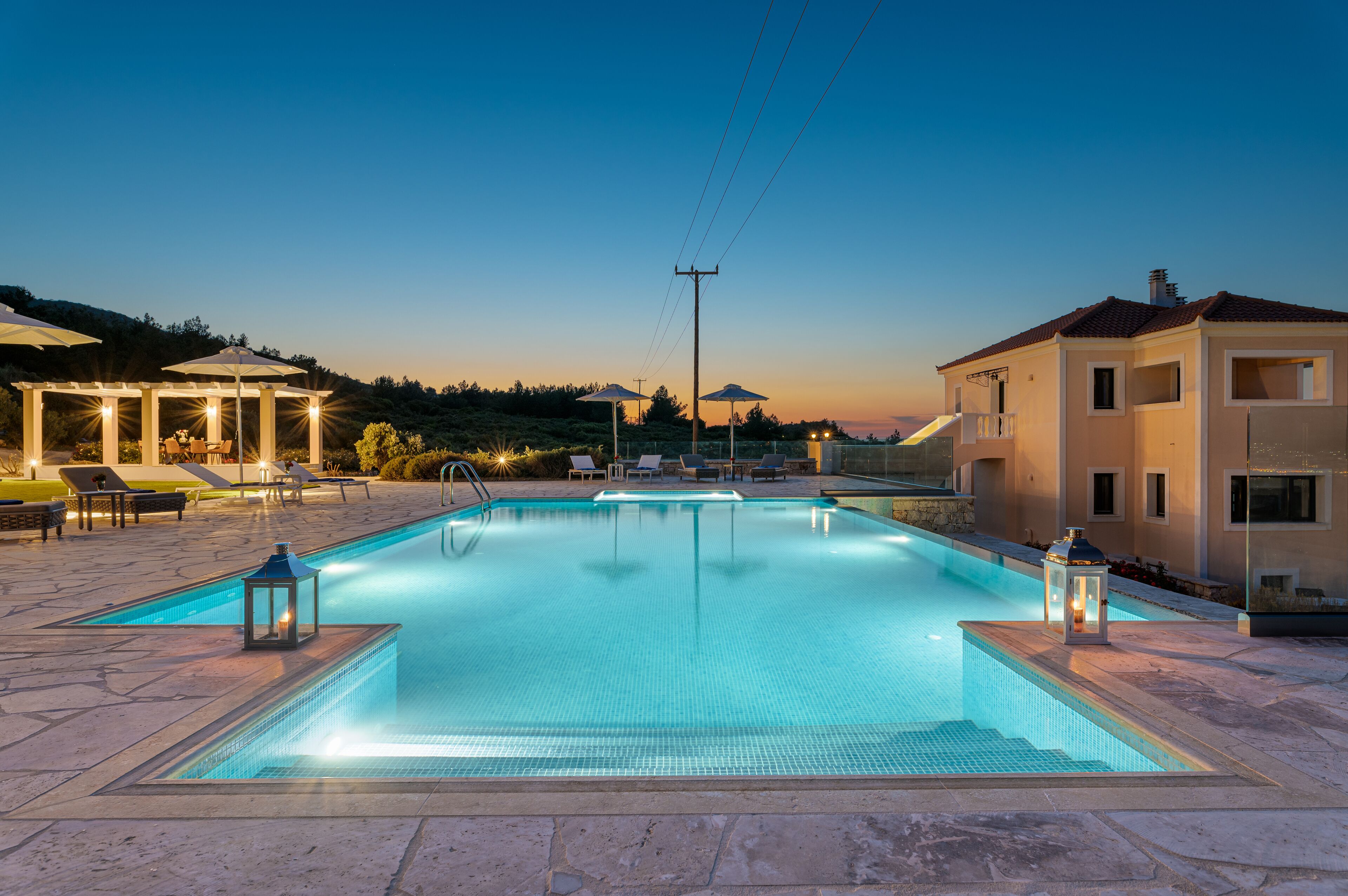 A rectangular swimming pool with a tiled edge, surrounded by lounge chairs and umbrellas, set against a backdrop of hills and a clear sky.