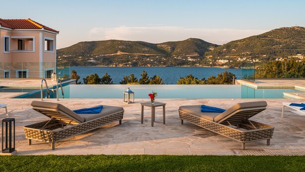 A poolside area with lounge chairs and a small table, overlooking a body of water and hills. - Grand View Villas (Samos)