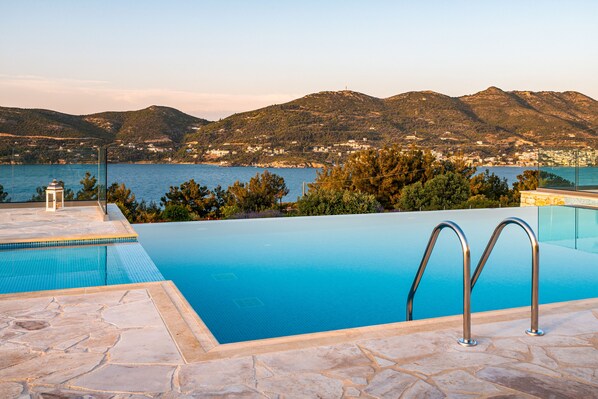 Una piscina con agua azul cristalina, rodeada de baldosas de piedra, con vistas a un paisaje montañoso y al mar.