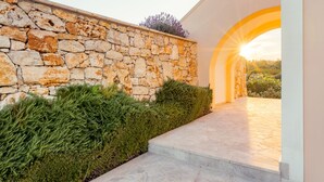 A stone wall with a green hedge and a stone pathway leading to an arched entrance. - Grand View Villas (Samos)