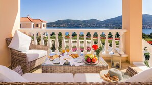 A balcony with wicker furniture, a dining table with breakfast items, and a view of mountains and water. - Grand View Villas (Samos)
