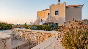 A modern building with a stone staircase leading to a white door, surrounded by lush greenery and flowering plants. - Grand View Villas (Samos)