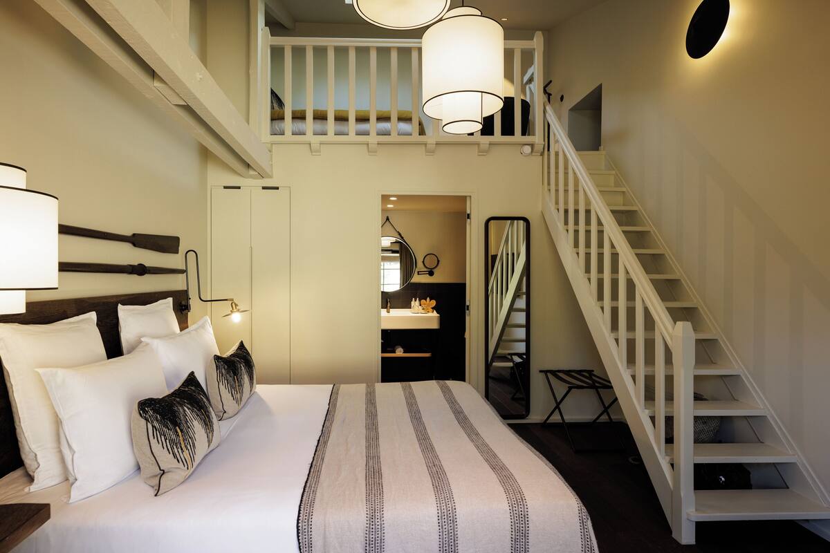 A bedroom with a large bed, a white staircase, a small bathroom, and a loft area with a balcony.