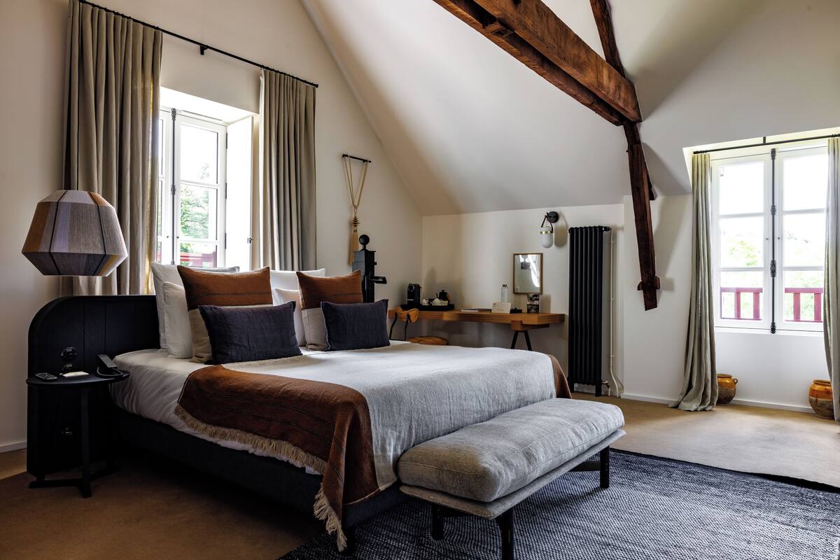 A bedroom with a large bed, a bedside table, a bench, a desk, and a window with curtains.