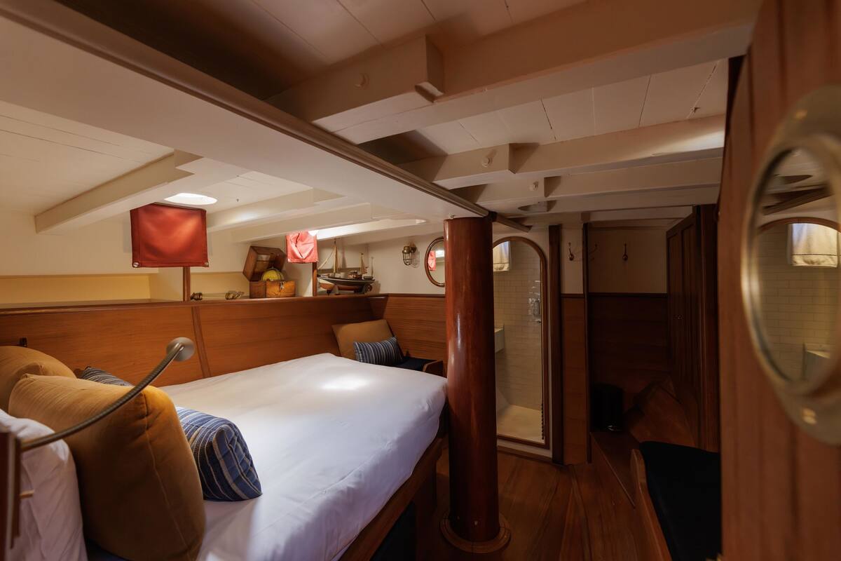 A cozy cabin with a wooden headboard, a bed with white linens, a small table with a lamp, and a window with curtains.
