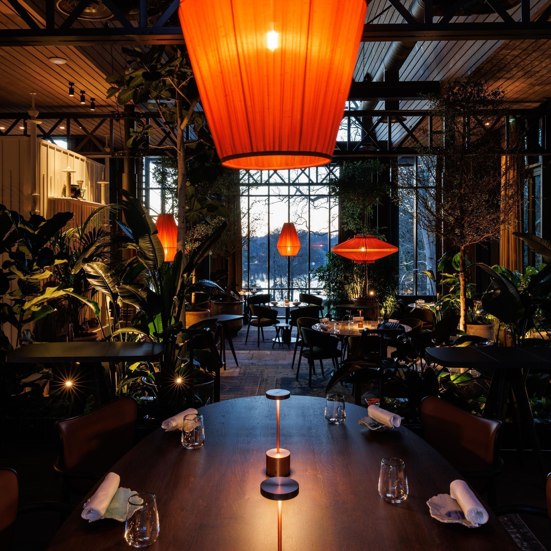 En restaurant med runde borde, hængende orange lamper og store vinduer med udsigt over det fri.