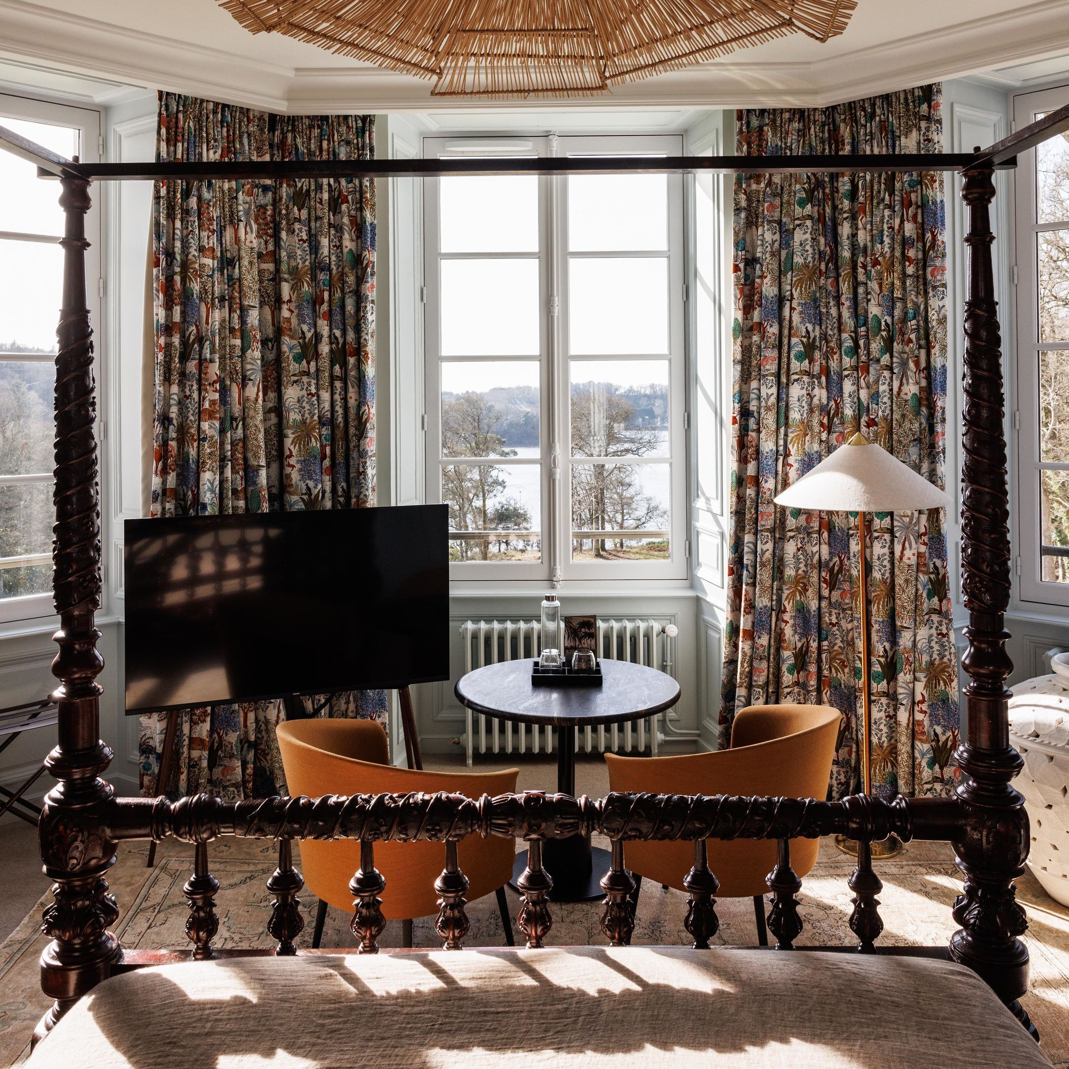A bedroom with a four-poster bed, a television, a table with chairs, and a view of a lake through large windows.