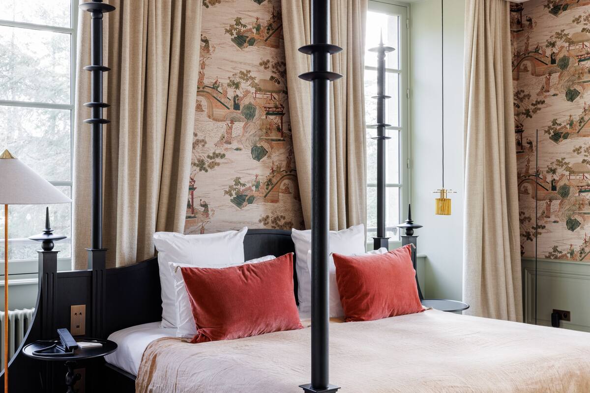 A hotel room with a large bed, red and white pillows, a black headboard, and a window with curtains.