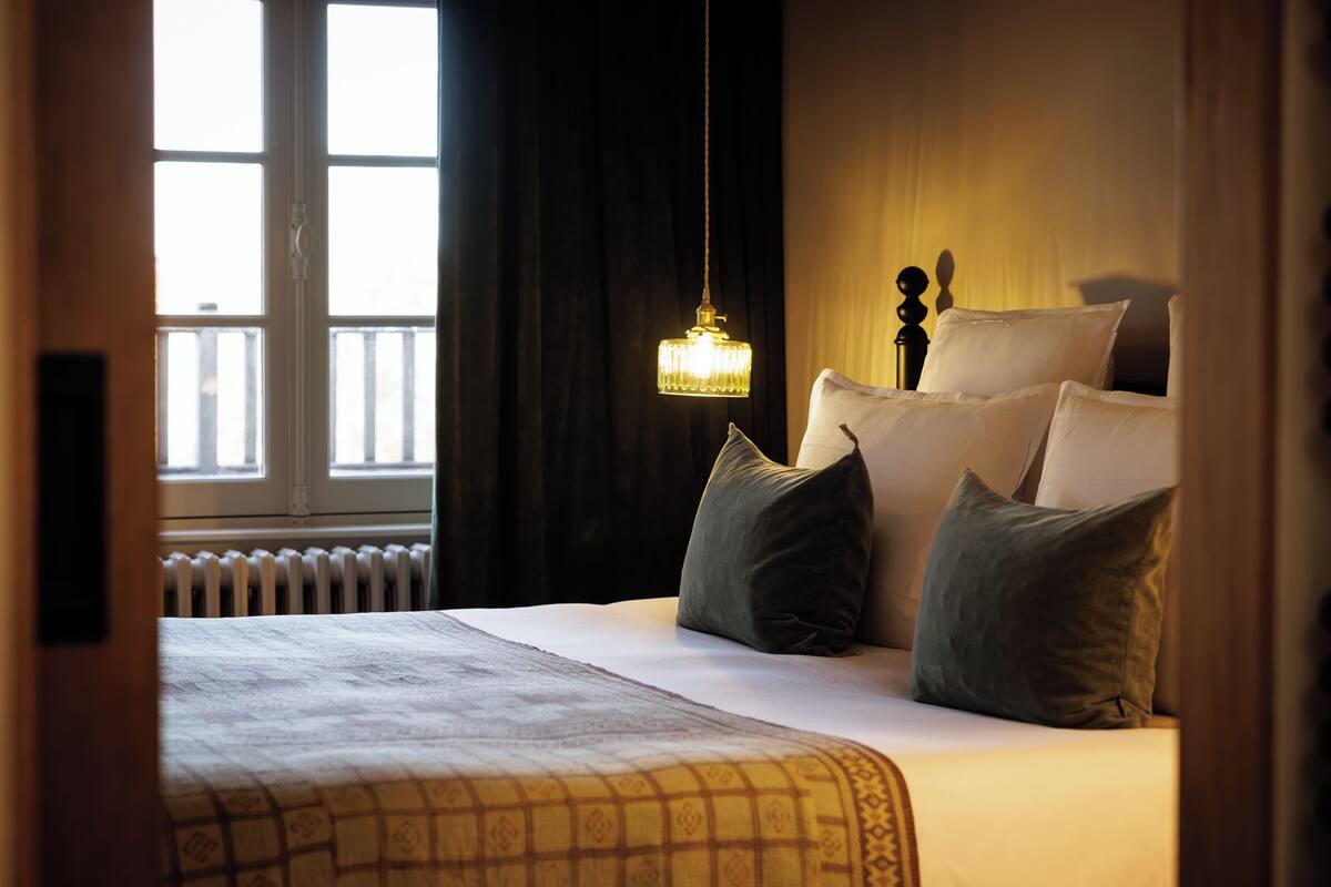 A neatly made bed with a patterned bedspread, a hanging light fixture, and a view of a window with curtains.