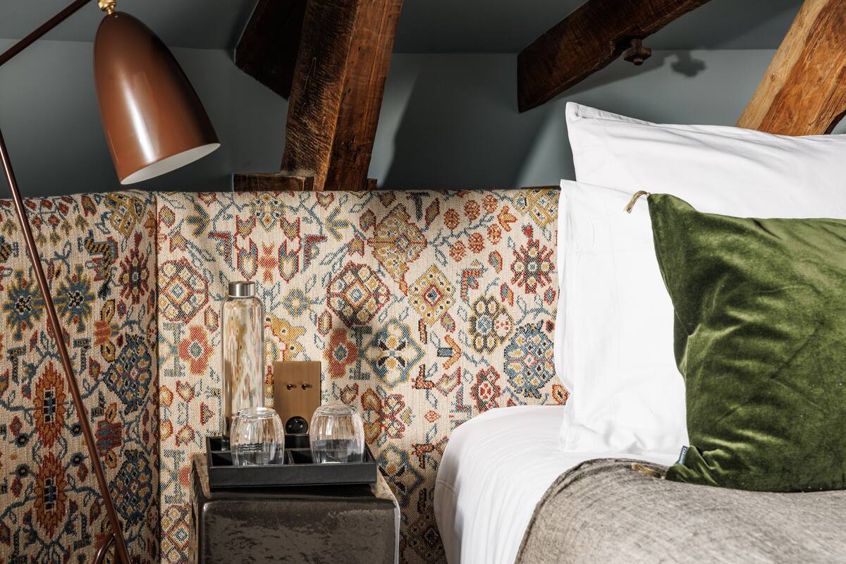 A bedroom with a patterned wall, a bed with white and green pillows, a bedside table with a lamp, and a wooden headboard.