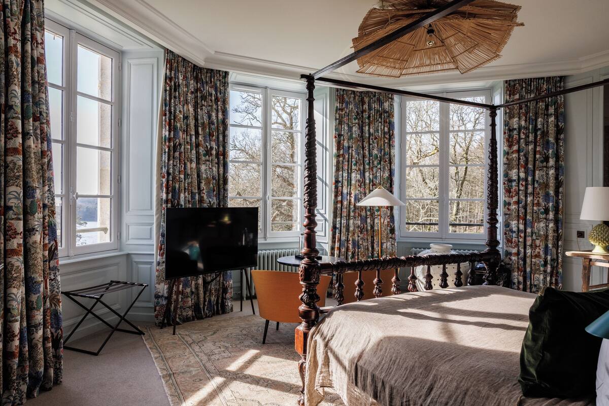 A four-poster bed with a canopy, a television, and a chair in a room with large windows and patterned curtains.