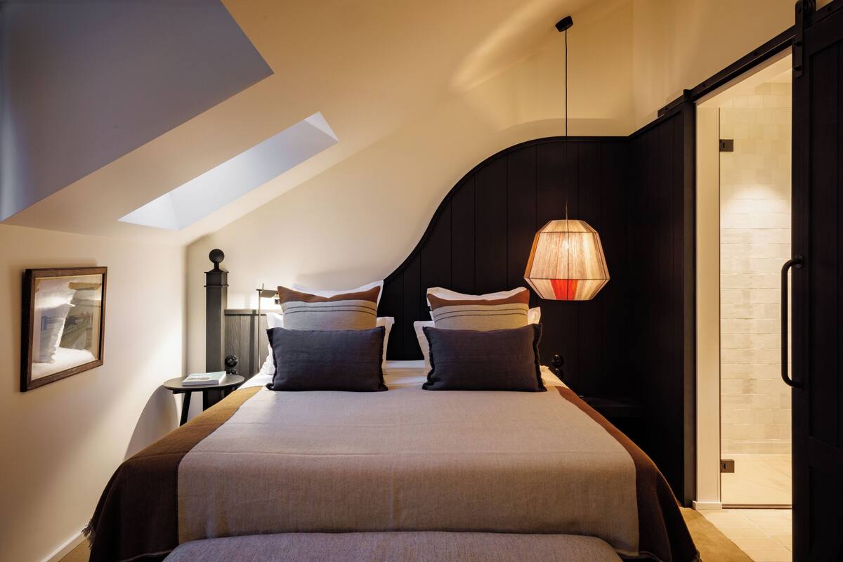 A bedroom with a large bed, a skylight, a bedside table, and a hanging light fixture.