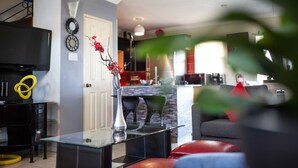 A modern living room with a glass coffee table, a flat-screen TV, and a dining area with a red chair. - The Oasis (Portmore)