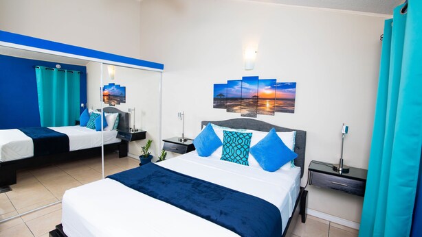 A hotel room with two beds, a large mirror, a sunset painting, and a teal curtain. - The Oasis (Portmore)