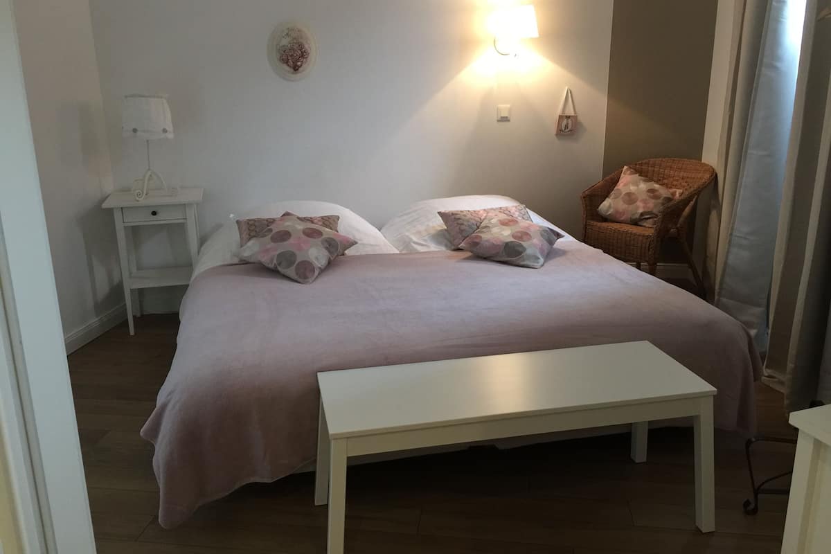 A bedroom with a bed, pillows, a bedside table, a lamp, a chair, and a wall-mounted light fixture.