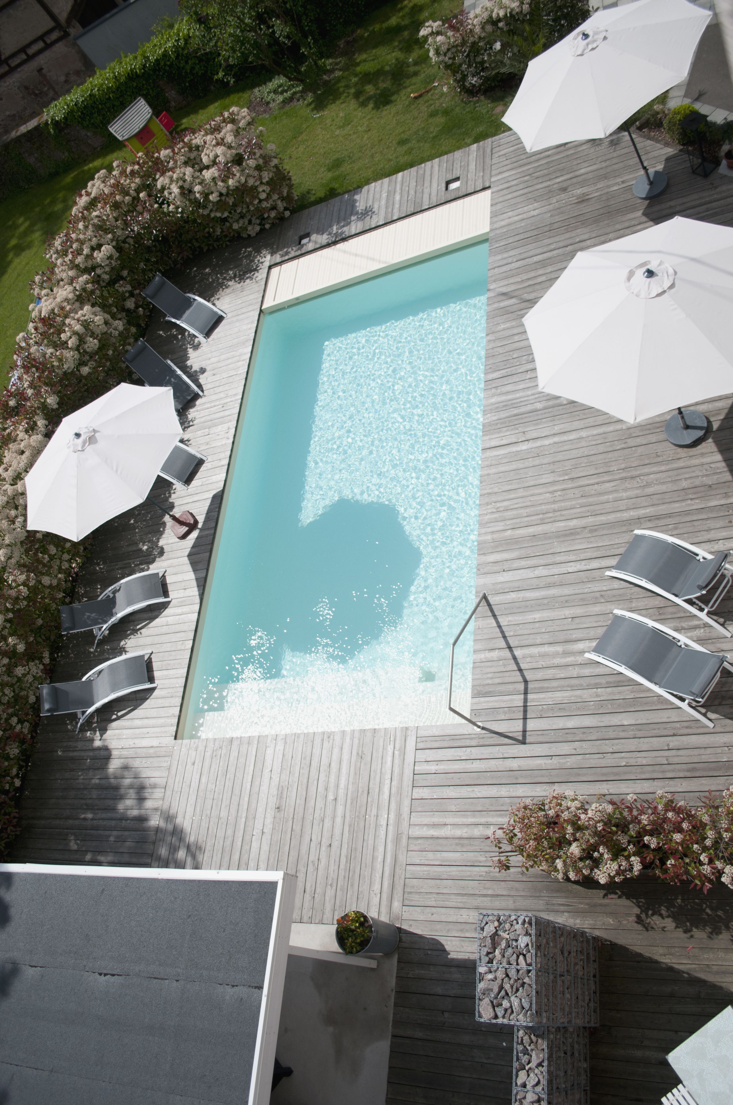 Vista aérea de una piscina rectangular rodeada por una terraza de madera, con sillones de descanso y sombrillas.