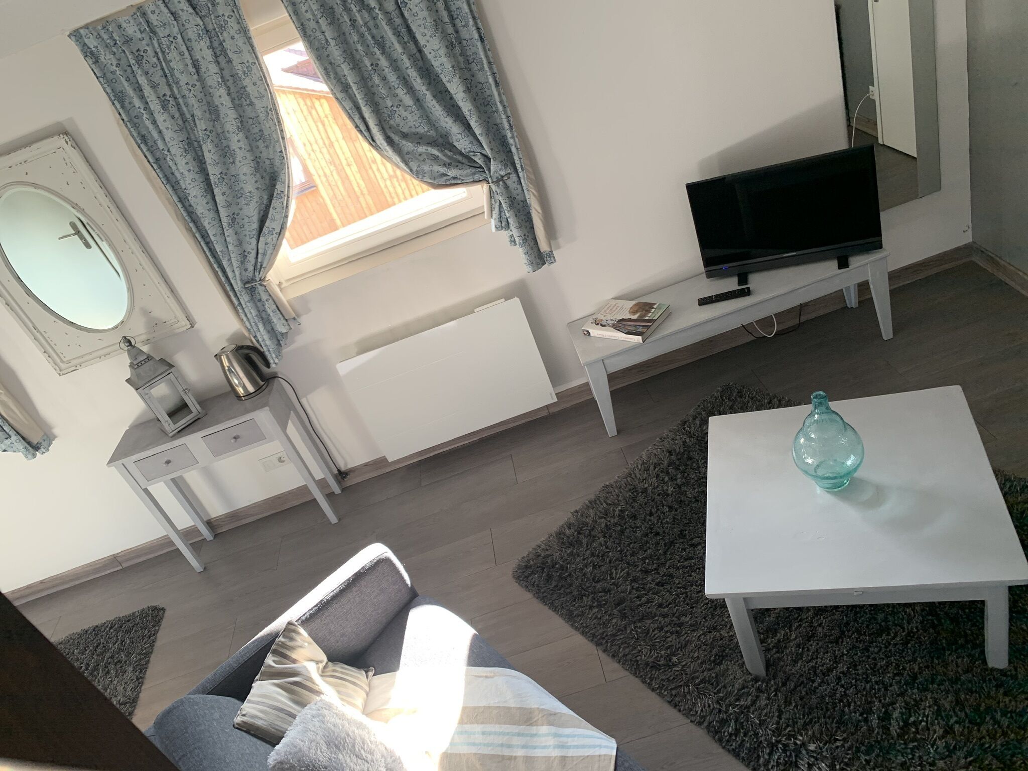 Sala de estar con sofá, mesa de centro, televisor sobre un mueble, una mesita con un libro y un control remoto, radiador, ventana con cortinas y espejo en la pared.