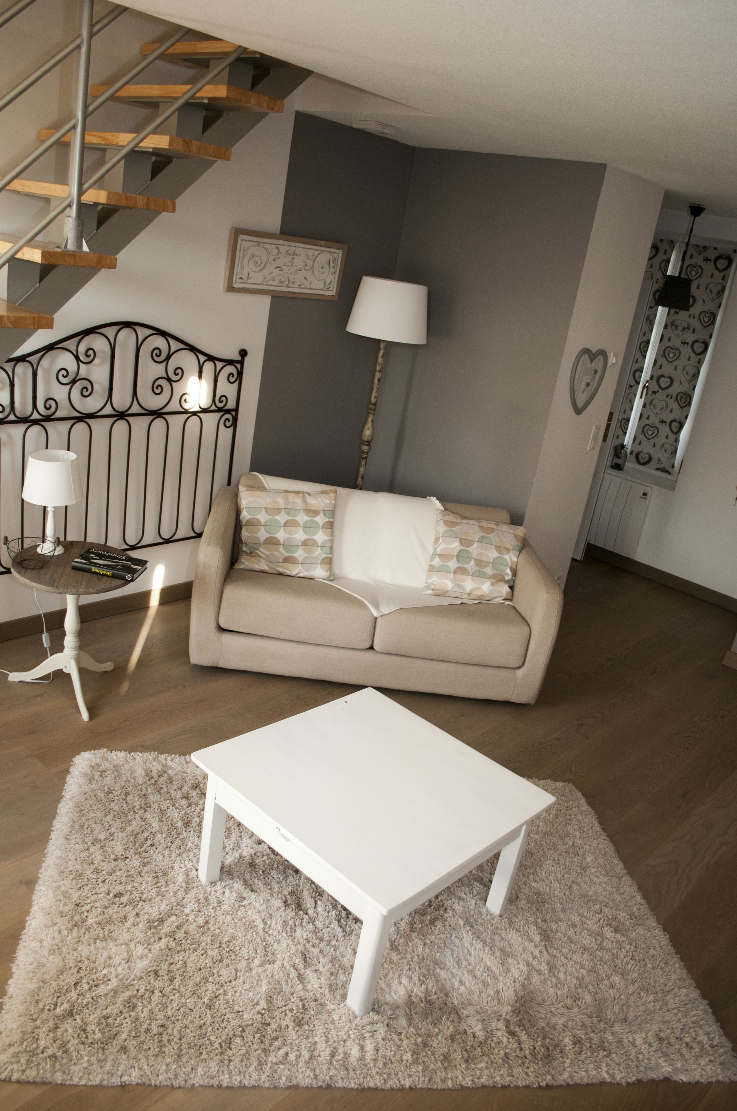A living room with a beige sofa, a white coffee table, a patterned rug, a metal staircase, and a wall-mounted lamp.