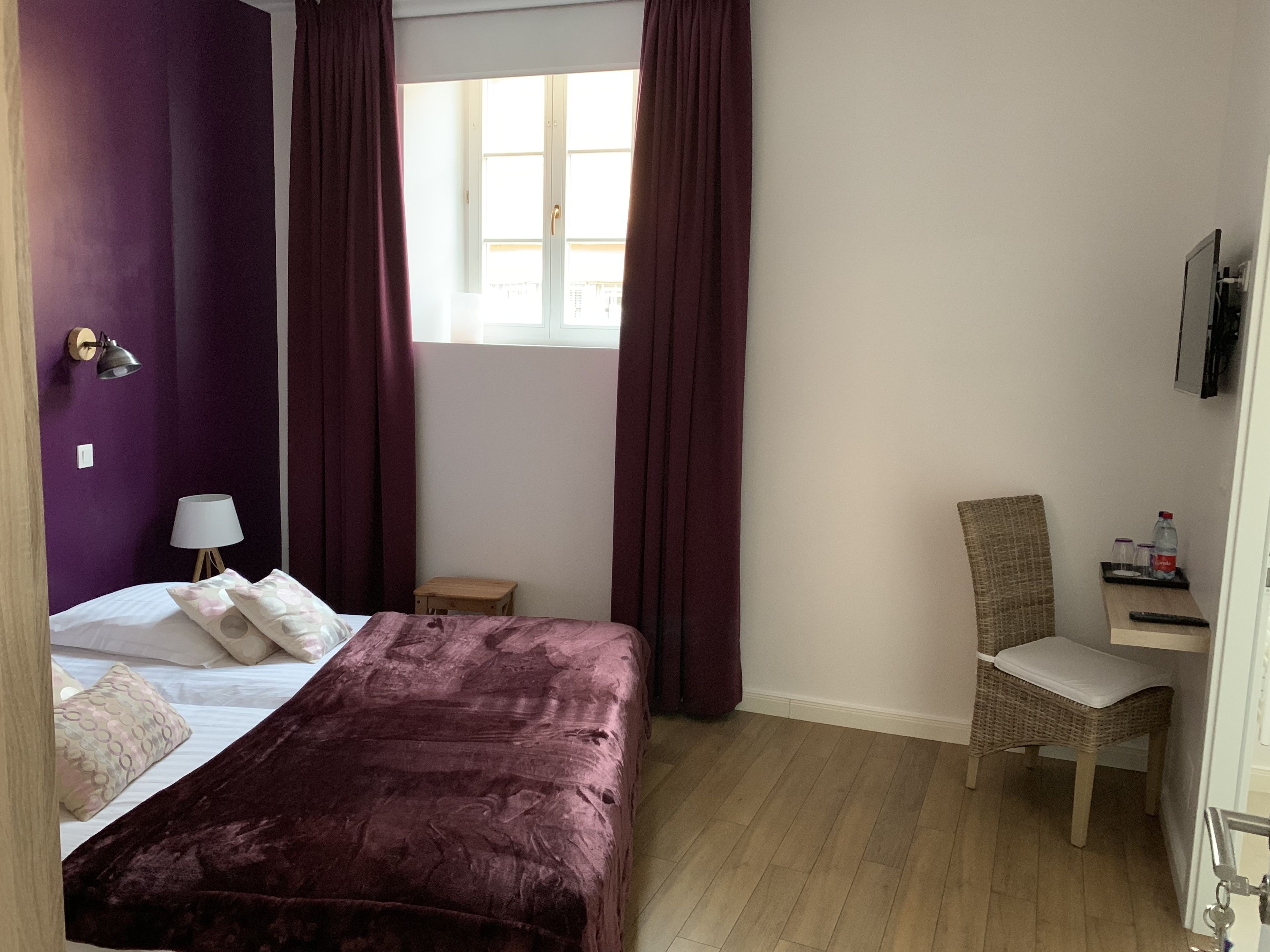 A hotel room with a bed, a small table, a chair, a window with curtains, and a wall-mounted TV.