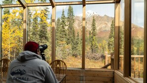 A person in a grey hoodie with a logo, sitting at a wooden table with a camera, overlooking a forest and mountains through large windows. - Wood River Lodge (Nenana)