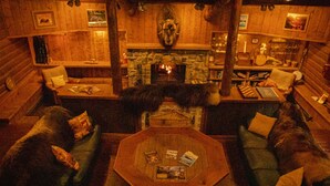 A cozy log cabin interior with a stone fireplace, wooden walls, and a large bear statue. There are two green sofas with pillows, a wooden coffee table, and a bookshelf filled with books and decorative items. - Wood River Lodge (Nenana)