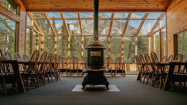 A room with a central stove, wooden chairs arranged around it, and large windows offering a mountain view. - Wood River Lodge (Nenana)