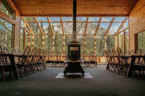 A room with a central stove, wooden chairs arranged around it, and large windows offering a mountain view. - Wood River Lodge (Nenana)