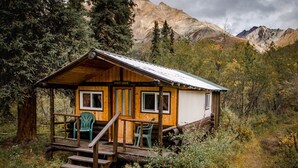 A rustic cabin named "Sheep Cabin" with a wooden exterior, a porch with benches, and a view of mountains in the background. - Wood River Lodge (Nenana)