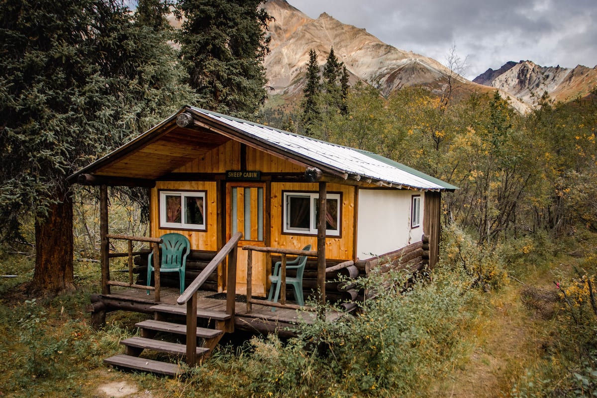 A rustic cabin named "Sheep Cabin" with a wooden exterior, a porch with benches, and a view of mountains in the background.