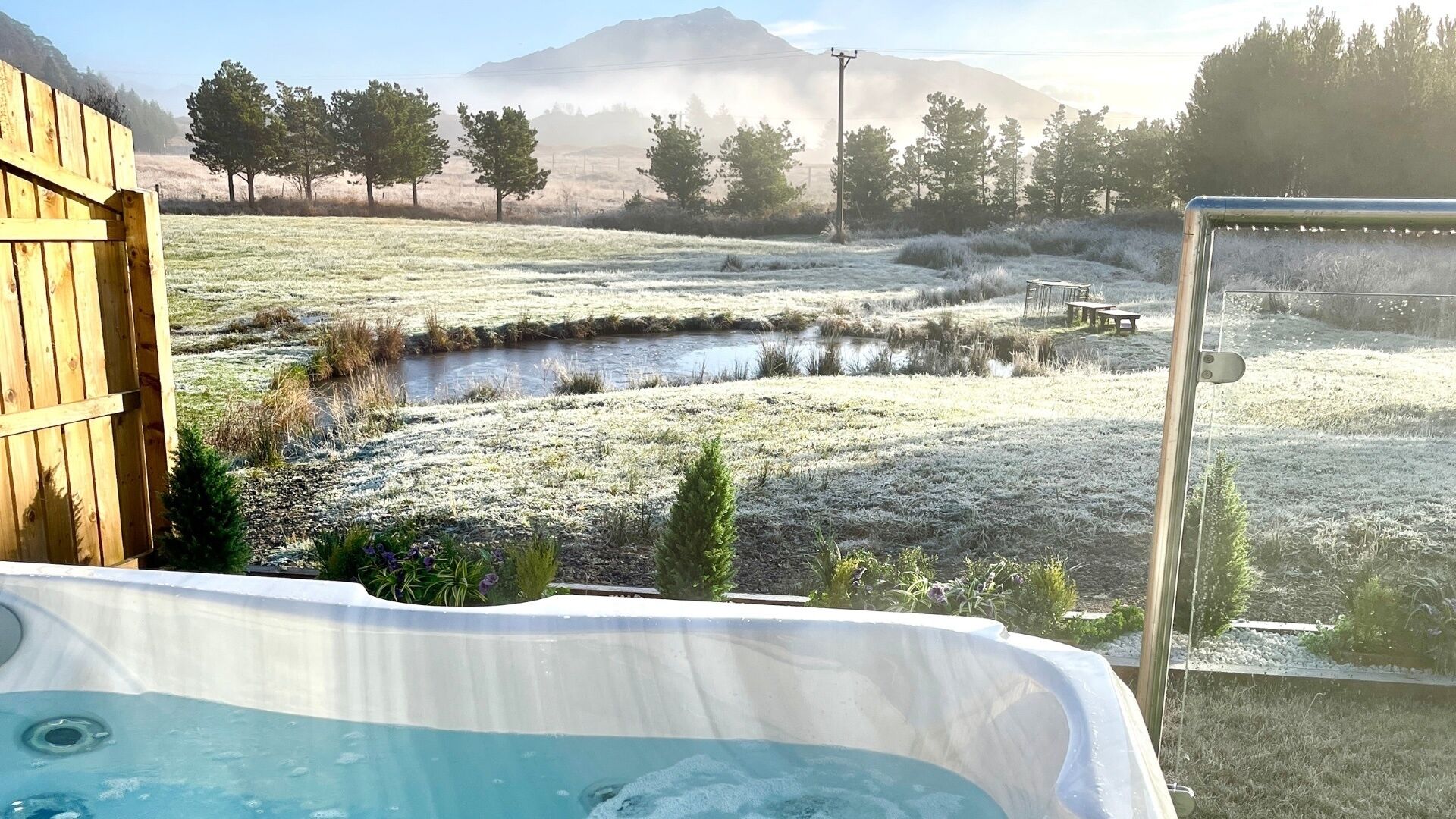 Un jacuzzi en un entorno natural con una terraza de madera, un pequeño estanque y una montaña al fondo.