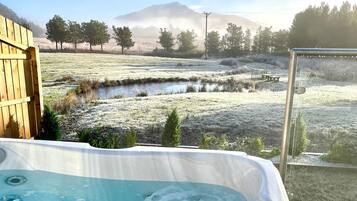 Un jacuzzi en un entorno natural con una terraza de madera, un pequeño estanque y una montaña al fondo.