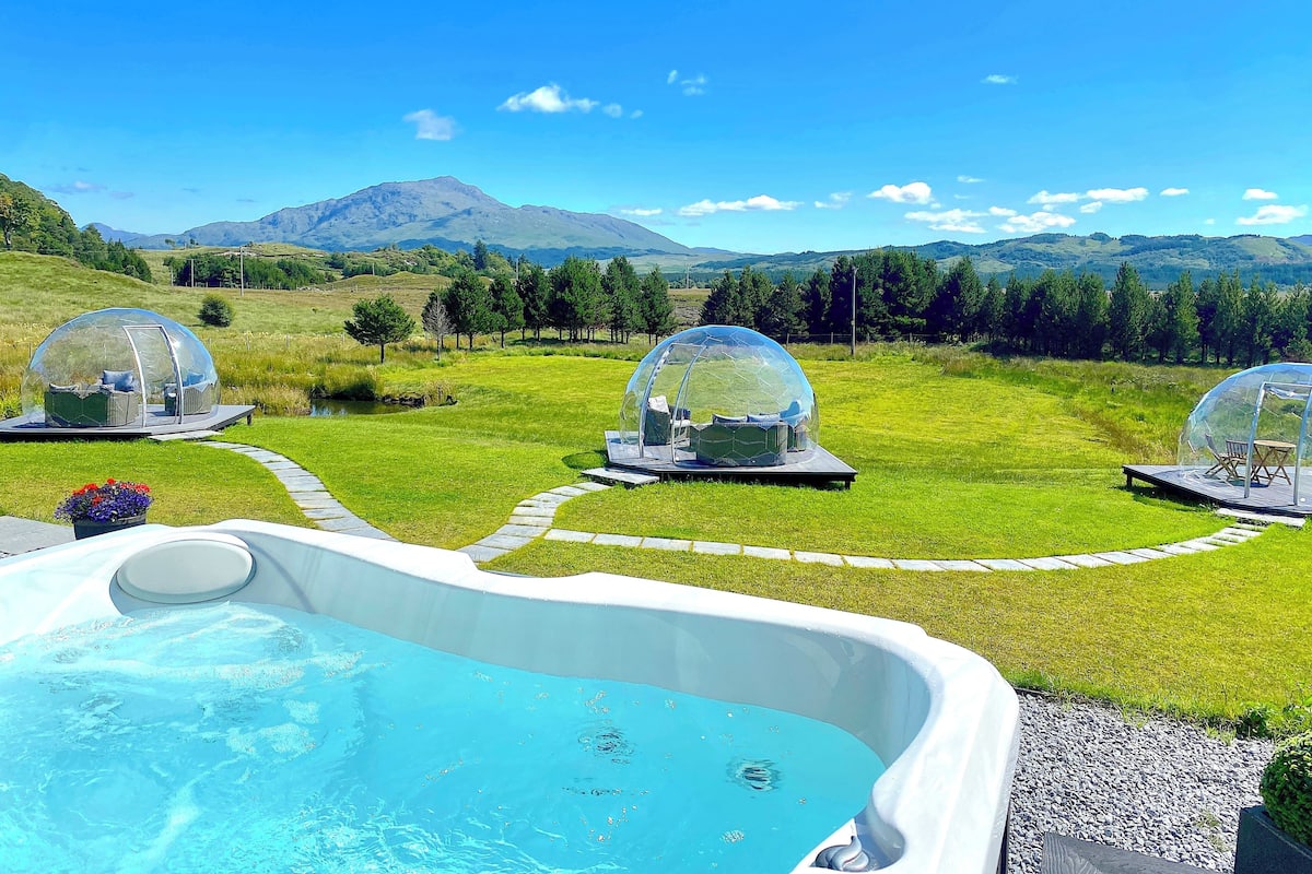 Ein Whirlpool mit drei transparenten Kuppeln in einer grünen Wiese, jeweils mit einer Sitzbank und einem Tisch.