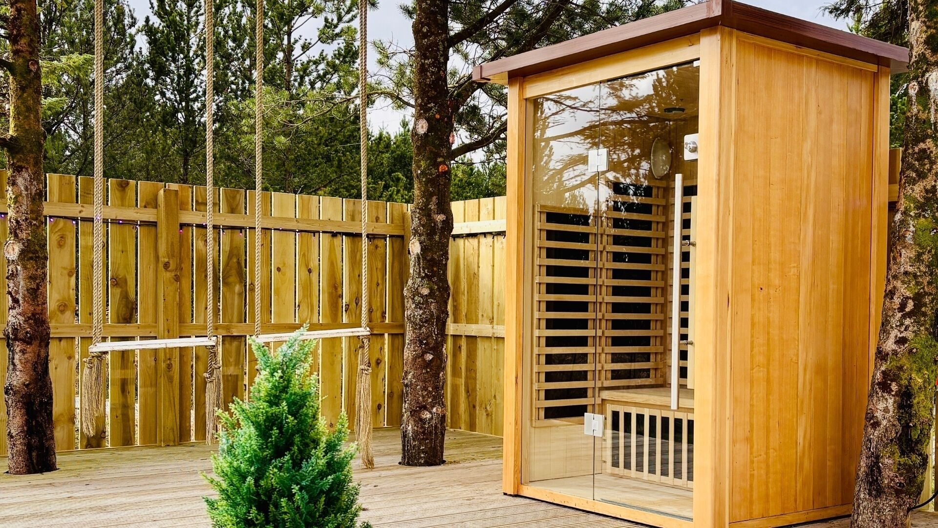 A wooden sauna structure with a glass door, surrounded by a wooden fence and trees.