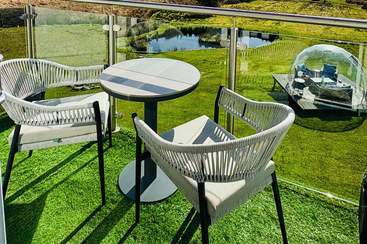 Ein Balkon mit zwei weißen Korbstühlen und einem runden Tisch, mit Blick auf eine Wiese mit Teich und entfernte Berge.