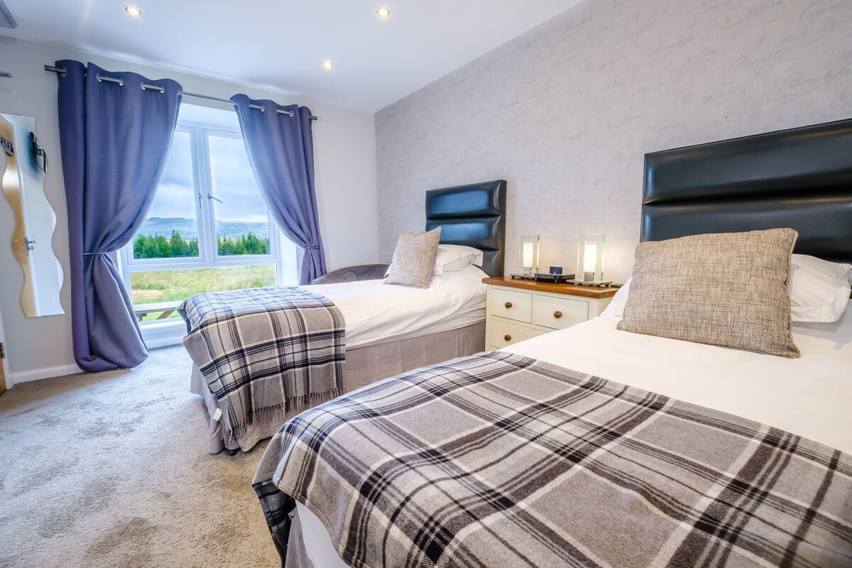 Ein Schlafzimmer mit zwei Betten, einem großen Fenster mit Vorhängen, einem Nachttisch mit Lampe und einem Kopfteil.
