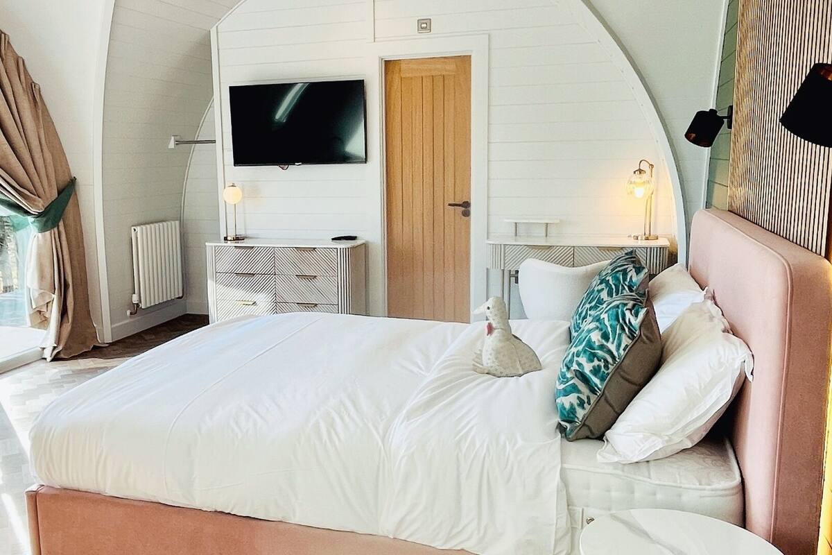 Ein Schlafzimmer mit einem großen Bett, einem an der Wand montierten Fernseher, einem Nachttisch und einem Fenster mit Vorhängen.