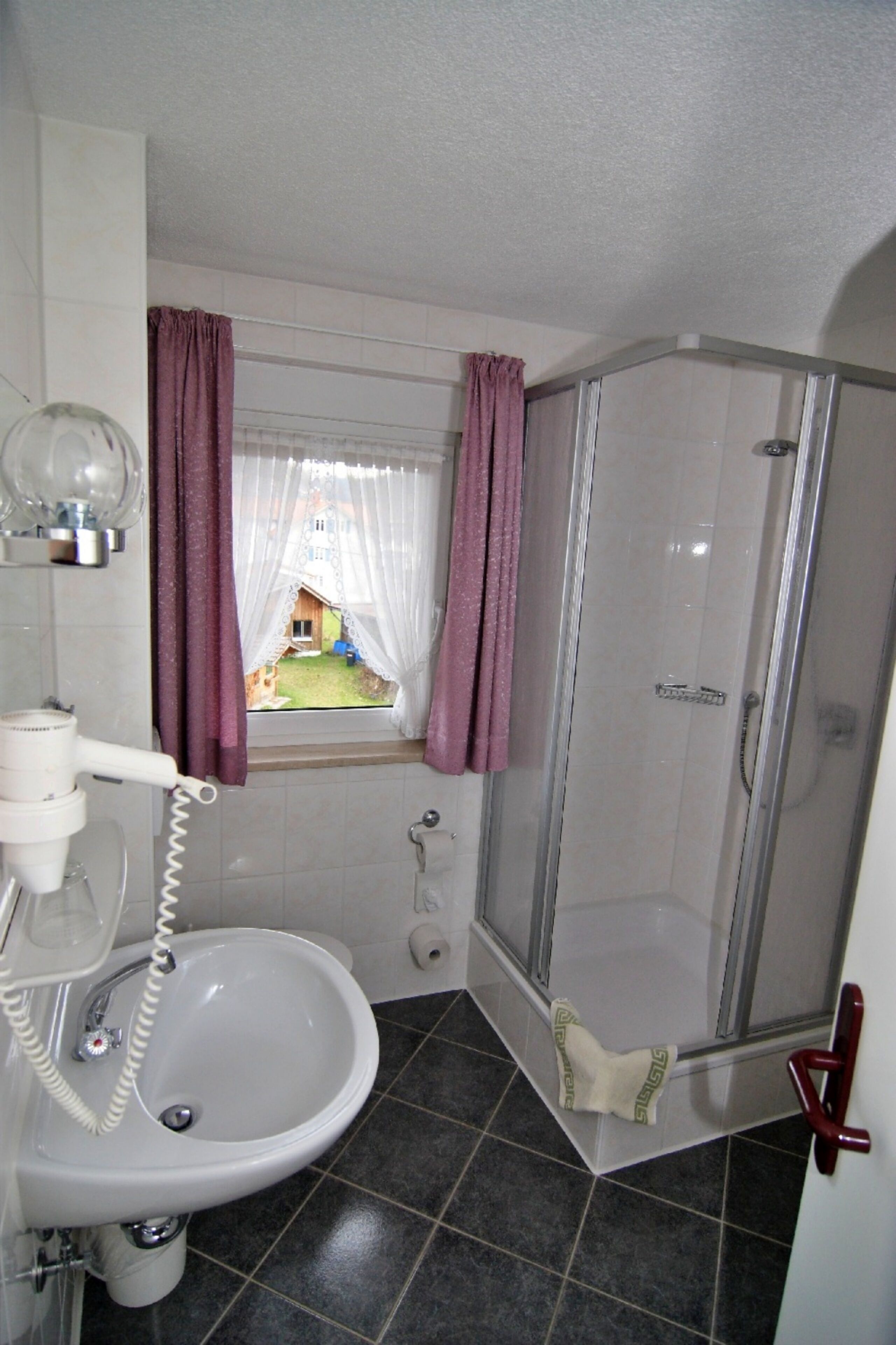 Un baño con lavamanos, secador de pelo, ducha y ventana con cortinas.