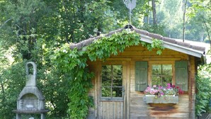 Una cabaña de madera rústica con chimenea, un comedero de pájaros de piedra y una caja de flores en la puerta.
