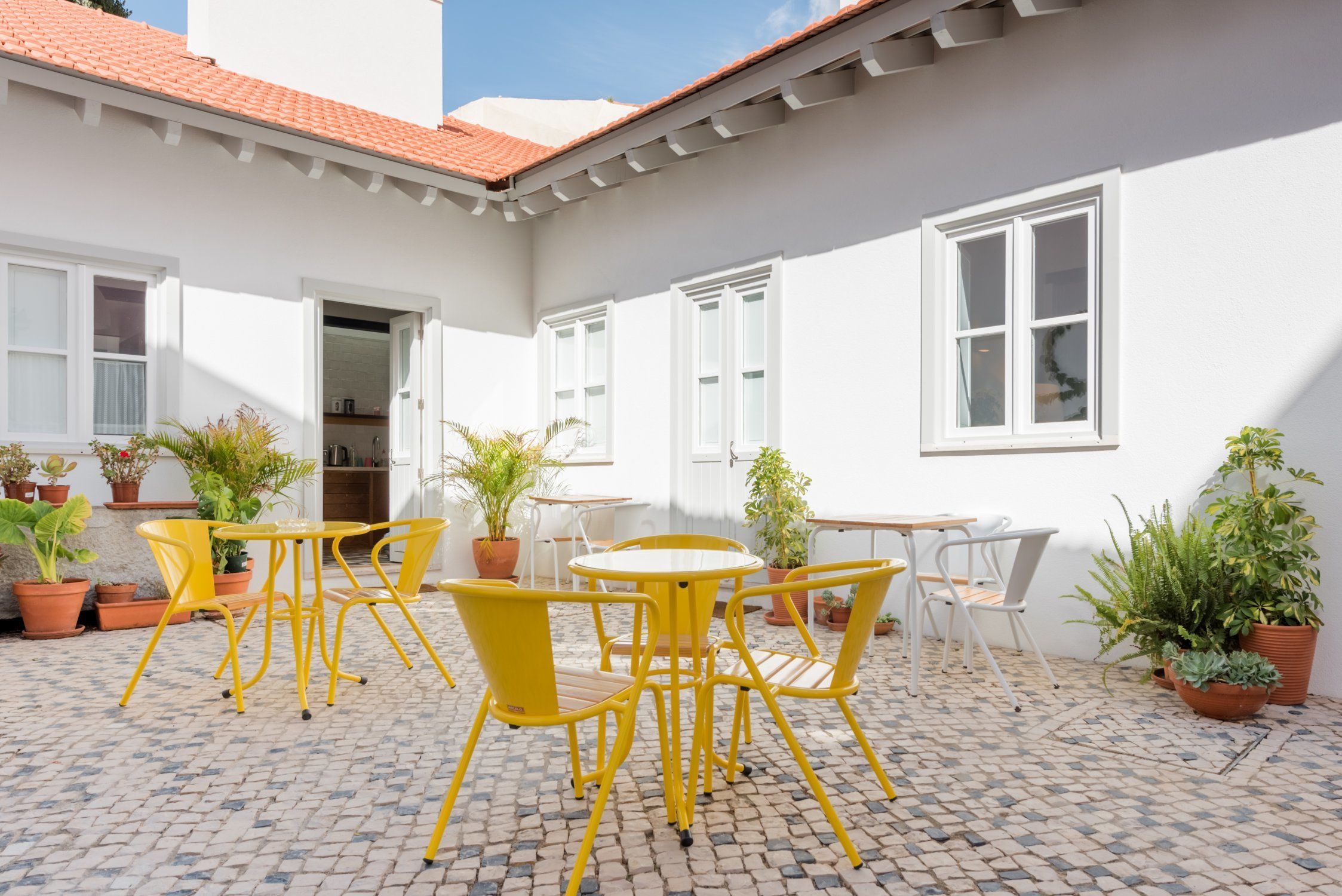 A courtyard with yellow metal chairs and tables, surrounded by potted plants and white walls.