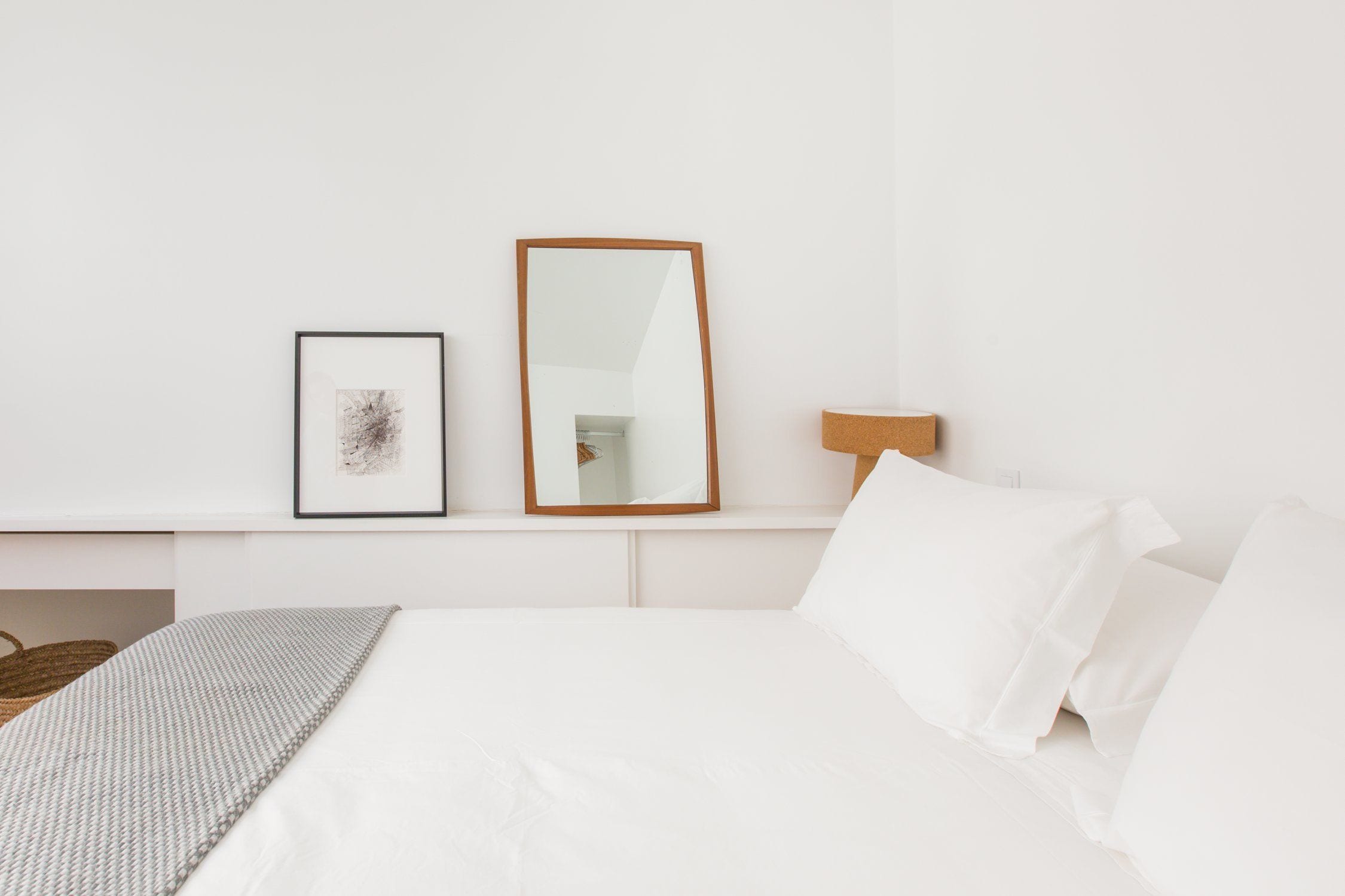 A neatly arranged bedroom with a bed, a framed picture, a mirror, and a bedside lamp.