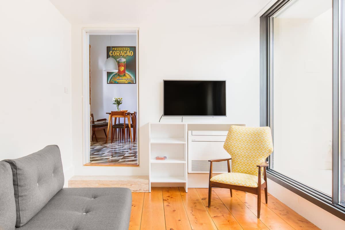 A modern living room with a grey sofa, a patterned armchair, a flat-screen TV on a white stand, and a wooden floor.
