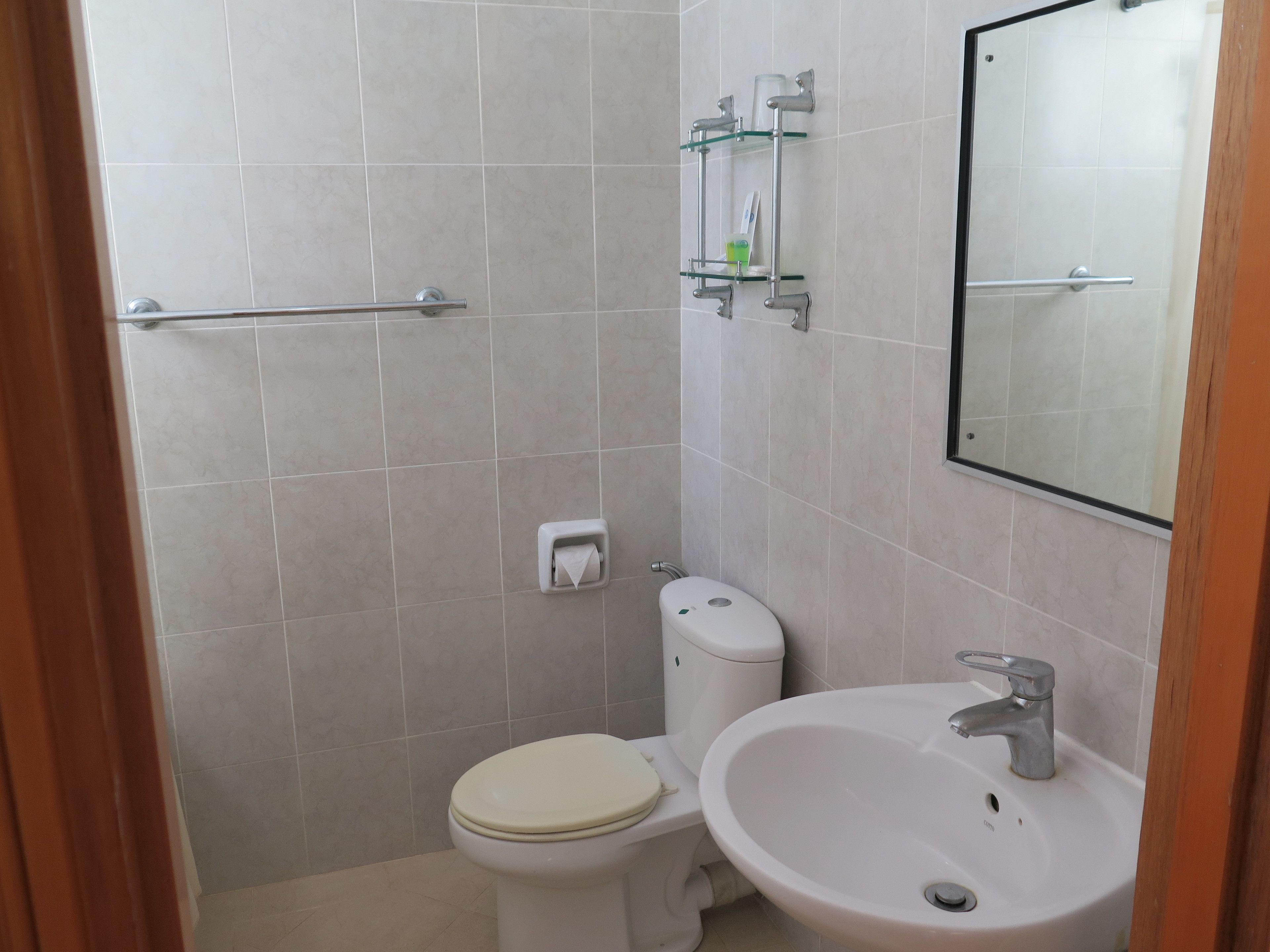 Une petite salle de bain propre, équipée de toilettes, d’un lavabo et d’une étagère en verre où sont rangés les articles de toilette.